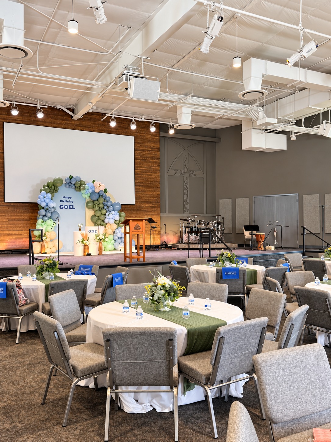Church first-birthday celebration with round tables, white tablecloths, sage cheesecloth runners and a sage-and-blue balloon backdrop.