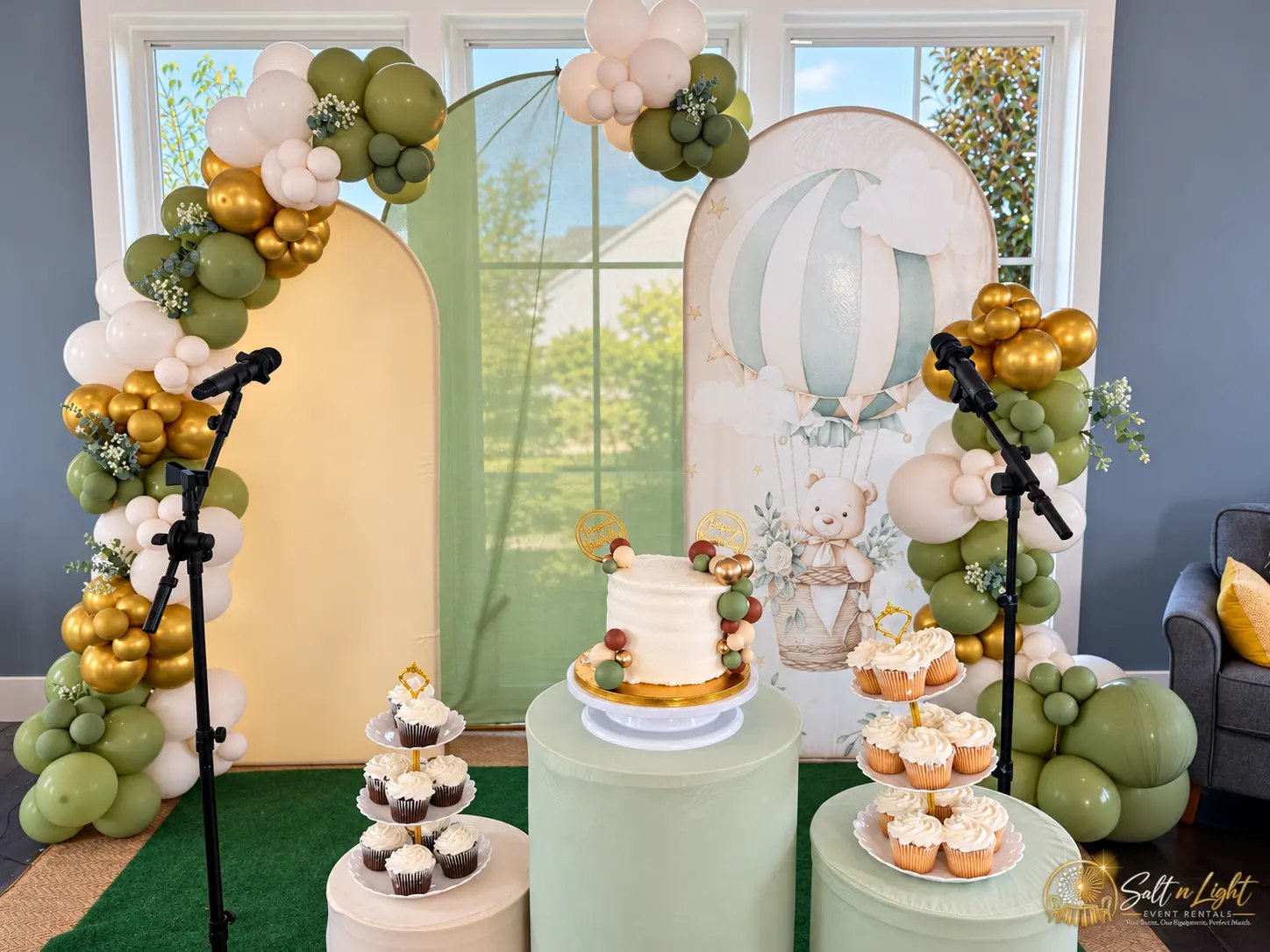 Sage green, cream and gold balloon backdrop with birthday cake, cupcakes and two microphones
