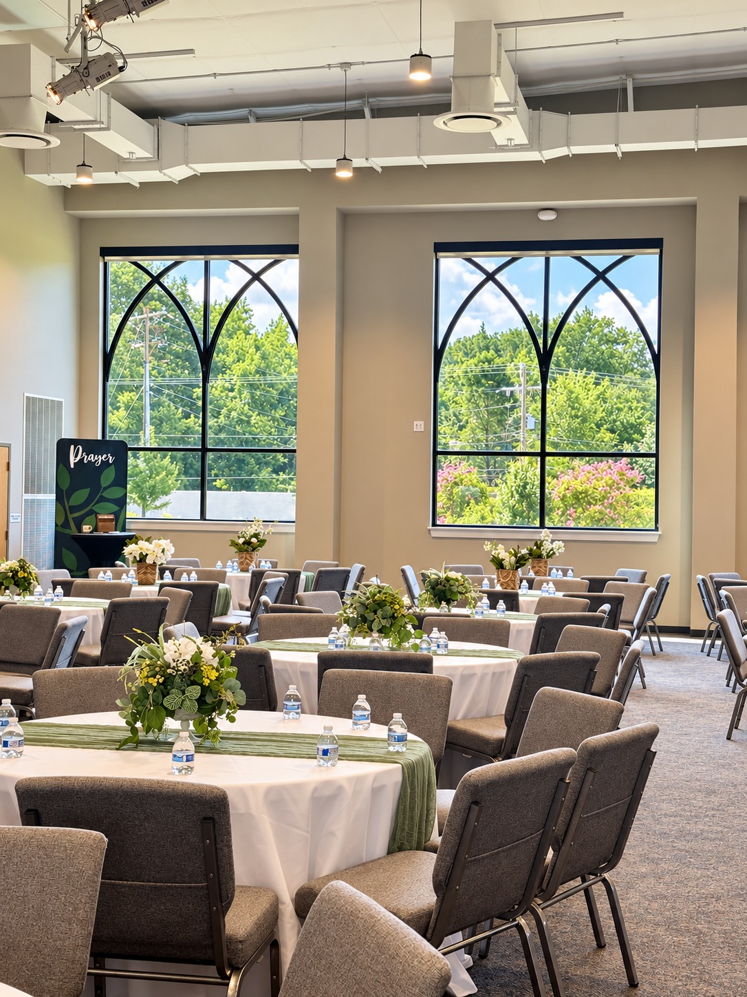 Round event tables dressed with white tablecloths, sage-green cheesecloth runners and floral centerpieces inside a church venue.